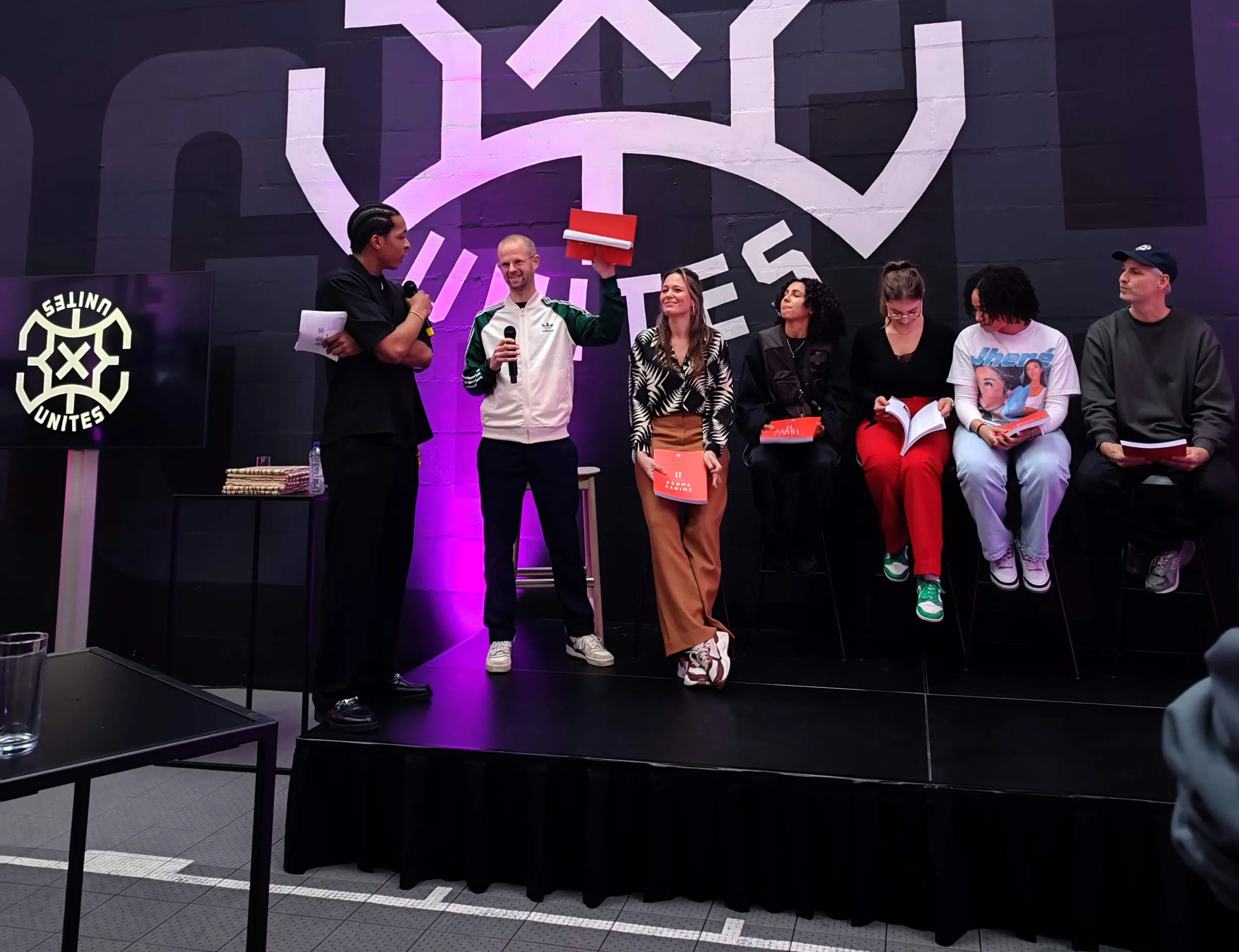 vijf mensen die op een podium zitten en een man die een rood boekje omhoog houd, ernaast een man met een microfoon. erachter het logo van 3X3 Unites
