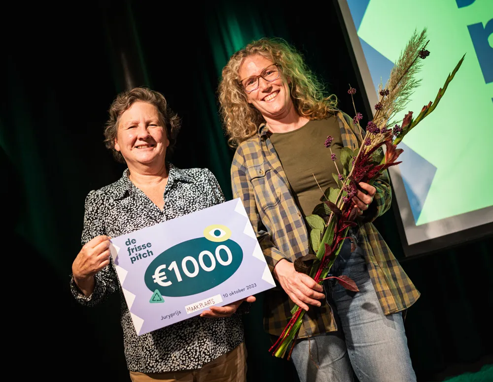 twee vrouwen op en podium met de ene geldcheque van 1000 euro, ernaast vrouw met een bosje droge bloemen