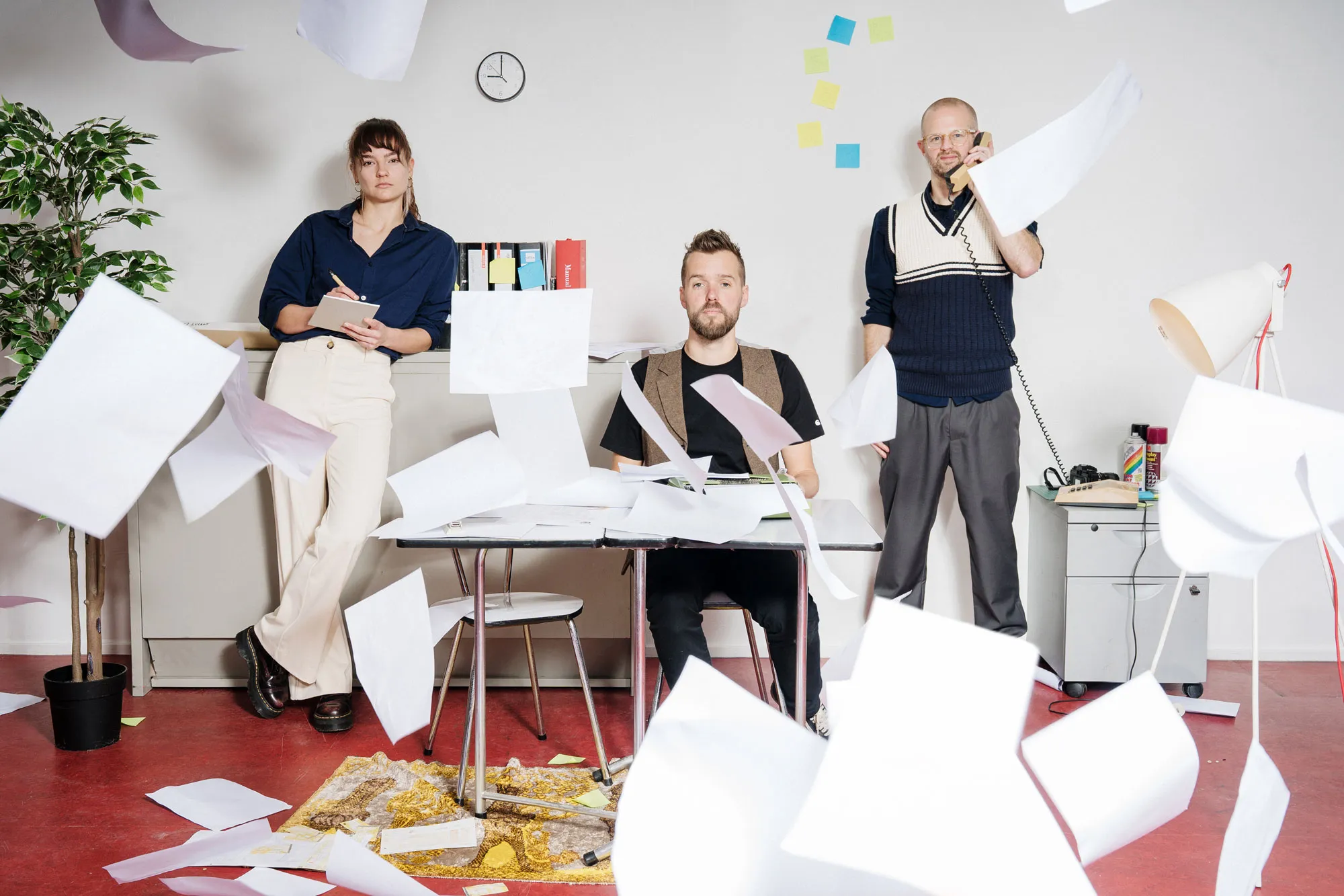 het team van Vrije Stijl in de studio, met papieren die door de lucht vliegen: een vrouw met een notitieblok, een man aan een bureau en een man met een telefoonhoorn